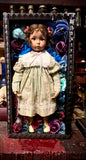 A vintage porcelain doll with brown curly hair and a white lace collar, wearing a pale green and pink striped dress, posed inside a black decorative frame filled with colorful fabric roses.