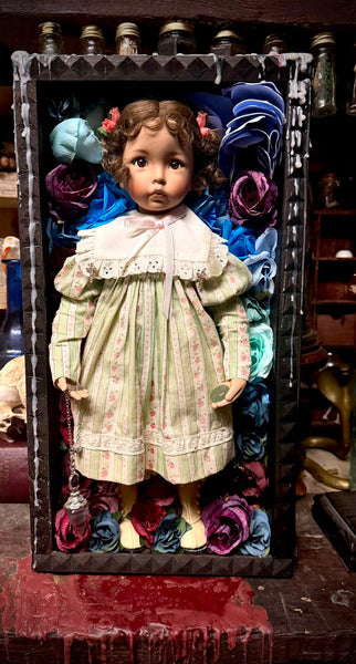 A vintage porcelain doll with brown curly hair and a white lace collar, wearing a pale green and pink striped dress, posed inside a black decorative frame filled with colorful fabric roses.